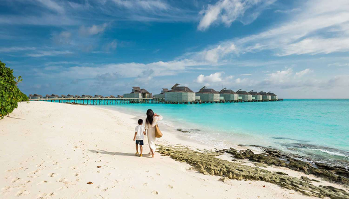 A mother and child walking on a white sand beach in the Maldives toward luxury overwater villas, representing a dream trip to avoid common travel mistakes.