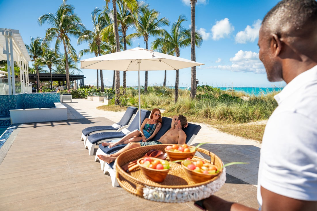 A couple relaxing on beach chairs at a luxury Maldives resort while a server brings fresh fruit snacks.