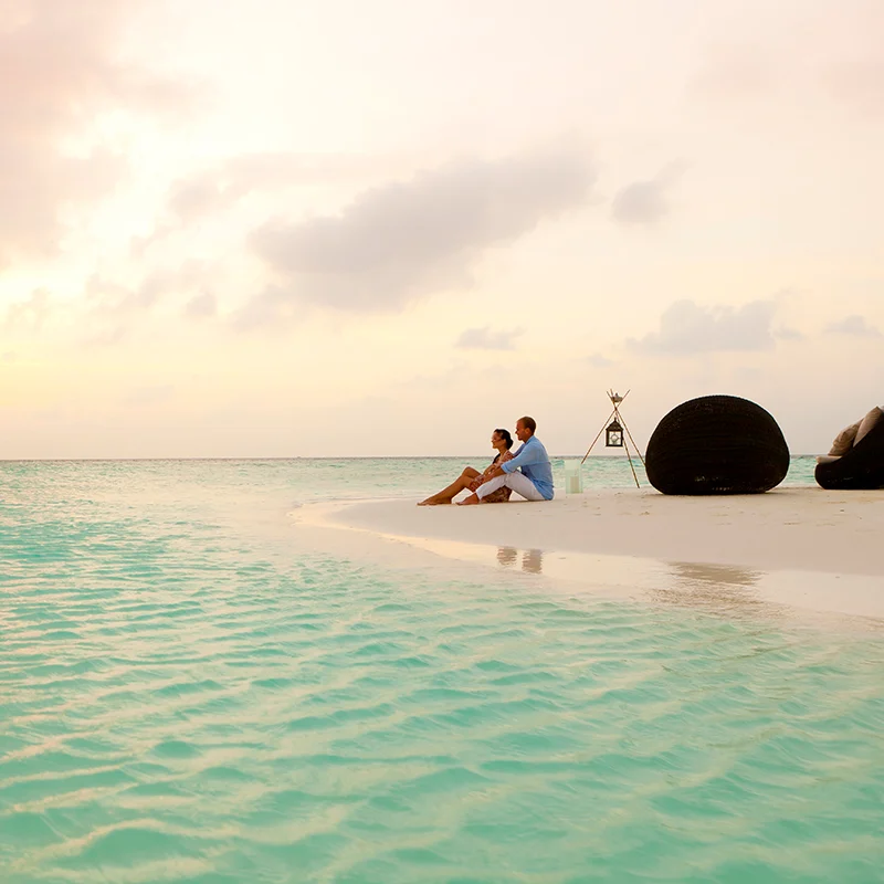 A romantic couple sitting on a white sandy beach at Baros Maldives during sunset, crystal clear turquoise water in the foreground.