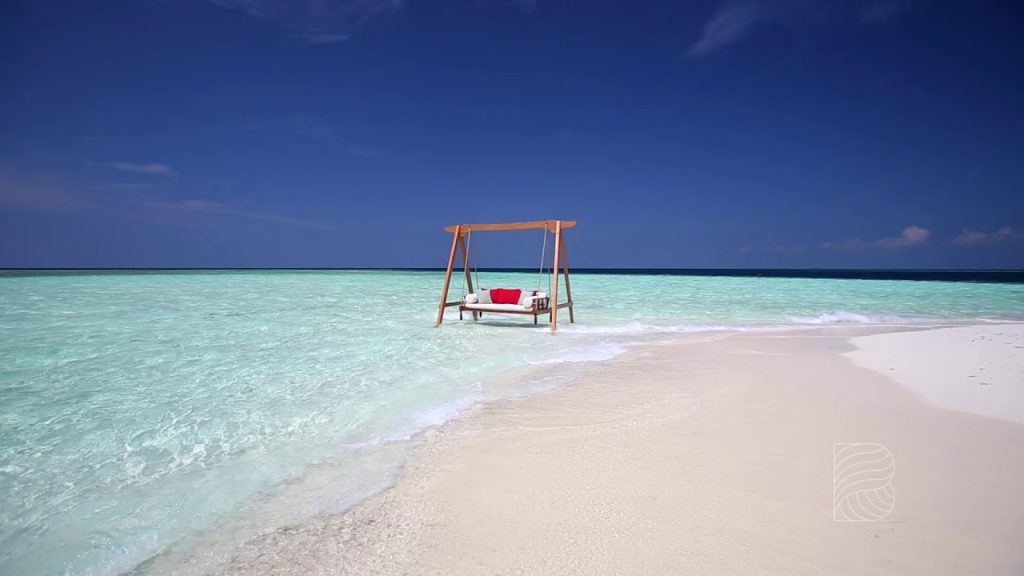 A luxury wooden swing with red pillows on a secluded white sandbank surrounded by crystal clear turquoise water at Baros Maldives resort.