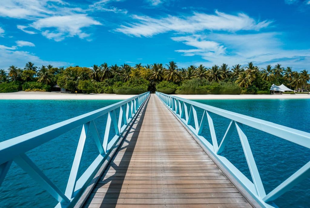 A long wooden bridge stretching over turquoise ocean water leading to a tropical island with palm trees at Conrad Maldives Rangali Island, a perfect instagrammable honeymoon spot.