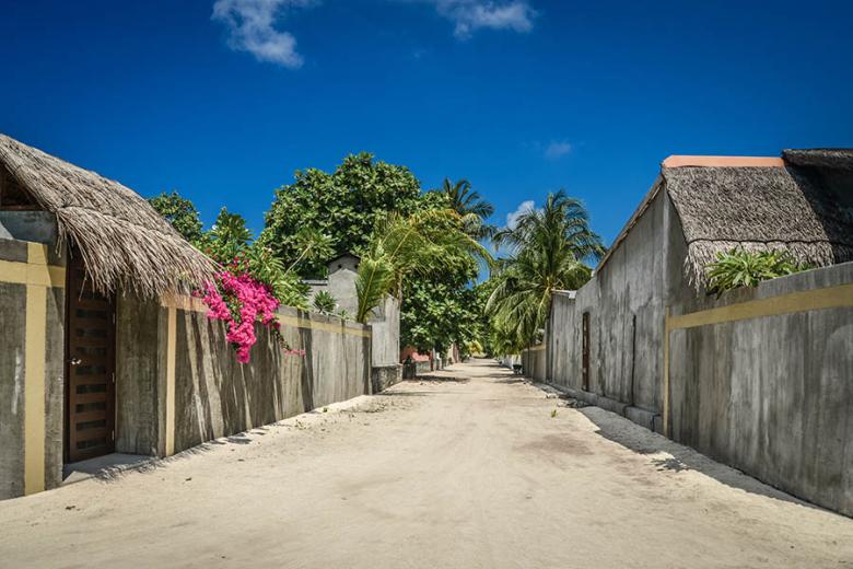 A sandy local street in Baa Atoll, Maldives, featuring traditional houses with thatched roofs and vibrant pink bougainvillea flowers under a clear blue sky.