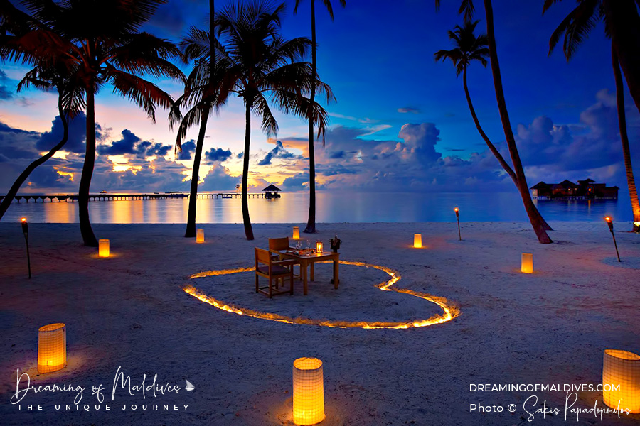 A romantic private candlelight dinner setup for two on a Maldives beach at sunset, featuring a heart-shaped light arrangement in the sand, palm trees, and ocean views.