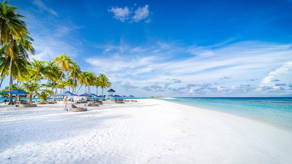 Pristine white sand beach and turquoise lagoon at Finolhu Baa Atoll, Maldives, with palm trees and beach umbrellas under a blue sky.