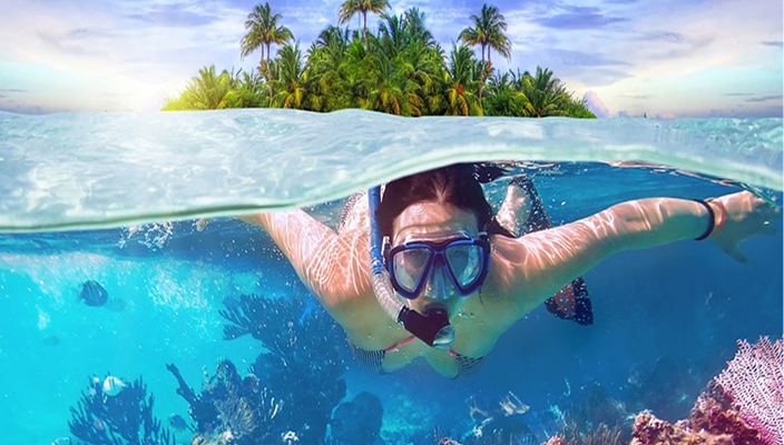 A woman snorkeling underwater in the Maldives with coral reefs and fish, featuring a tropical island with palm trees in the background.