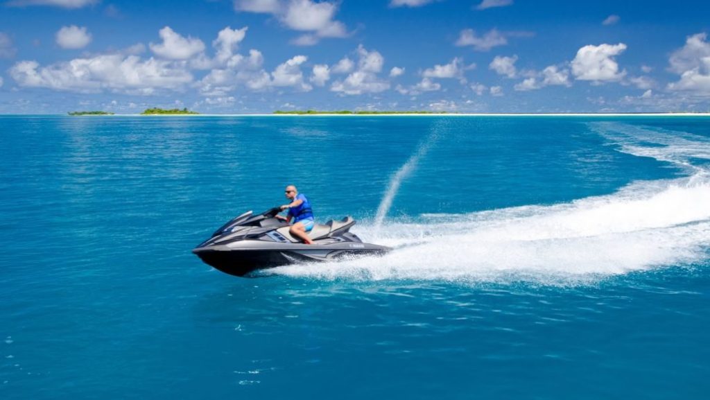 A man riding a grey Yamaha jet ski across the turquoise blue waters of the Maldives with a tropical island in the background.