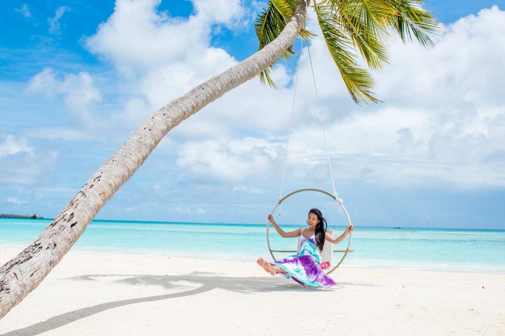 A woman in a purple dress sitting on a hanging ring swing attached to a leaning palm tree on a white sandy beach with turquoise ocean water at Kandima Maldives.