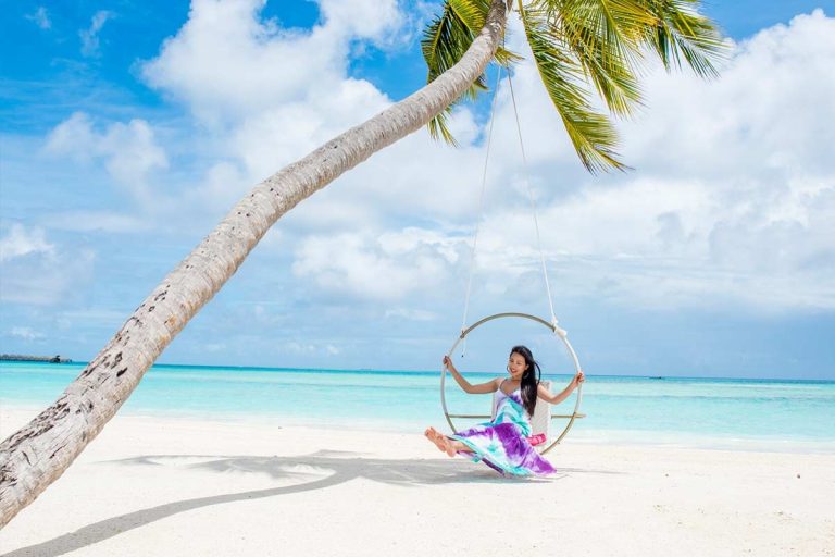 A woman in a purple dress sitting on a hanging ring swing attached to a leaning palm tree on a white sandy beach with turquoise ocean water at Kandima Maldives.