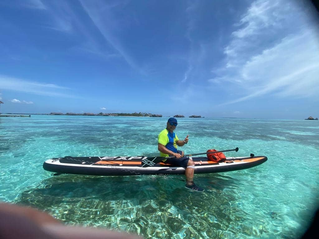 A man sitting on a stand-up paddleboard (SUP) in the turquoise lagoons of the Maldives under a bright blue sky, showcasing water sports adventures.