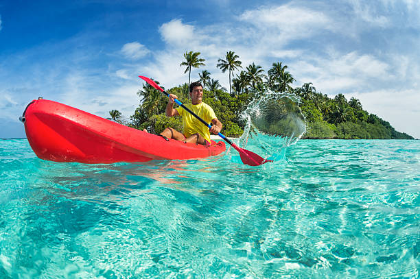 A man in a yellow shirt paddling a red kayak through turquoise ocean water near a tropical island with palm trees in the Maldives.