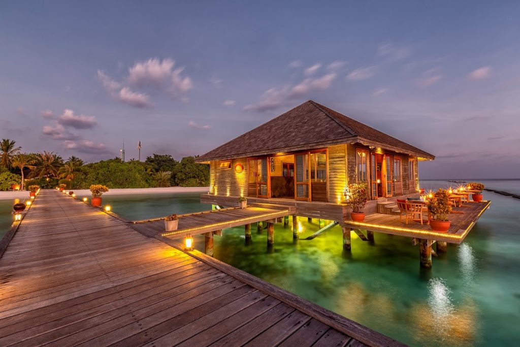 A luxury overwater wooden bar at Komandoo Island Resort Maldives during sunset with golden lights reflecting on turquoise water.