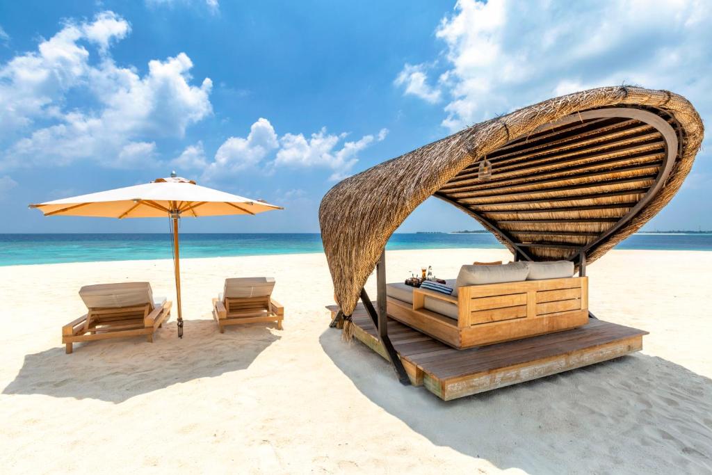 A luxury thatched-roof beach cabana with comfortable seating on the white sandy beach of Kudadoo Maldives Private Island, overlooking the turquoise ocean under a blue sky.
