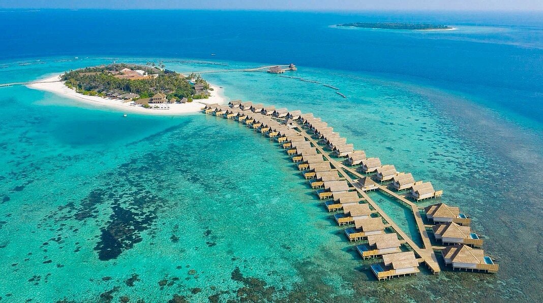 Aerial view of a luxury resort in Raa Atoll, Maldives, featuring a long wooden jetty connected to white overwater villas with private pools and thatched roofs in a turquoise lagoon.