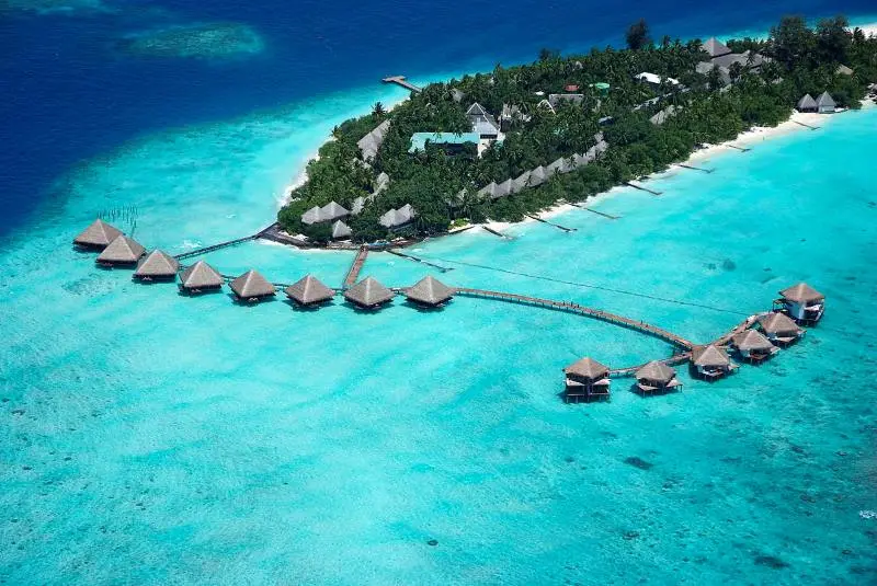 Aerial view of a luxury honeymoon resort in Maafushi Island, Maldives, featuring turquoise ocean water and overwater wooden villas.