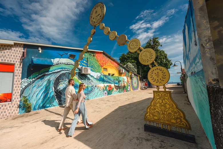 Two tourists walking past a colorful mural and gold traditional archway on a local Maldivian island.