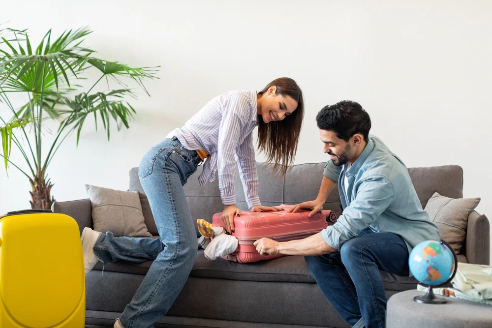 A couple struggling to close an overpacked pink suitcase on a sofa, illustrating common travel packing mistakes to avoid for a Maldives vacation.