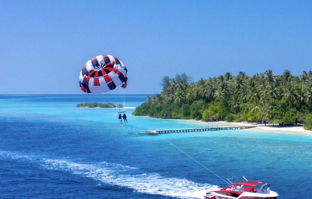 Two people parasailing high above a tropical island with white sand beaches and palm trees in the Maldives.