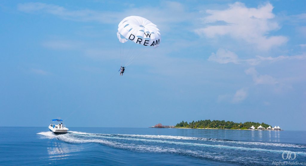 A person parasailing over the crystal clear turquoise waters of the Maldives with a tropical island and a speedboat in the background.