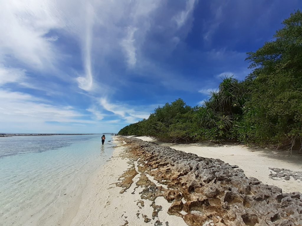 A woman walking along a pristine white sand beach with turquoise water and lush tropical greenery in Lhaviyani Atoll, Maldives.