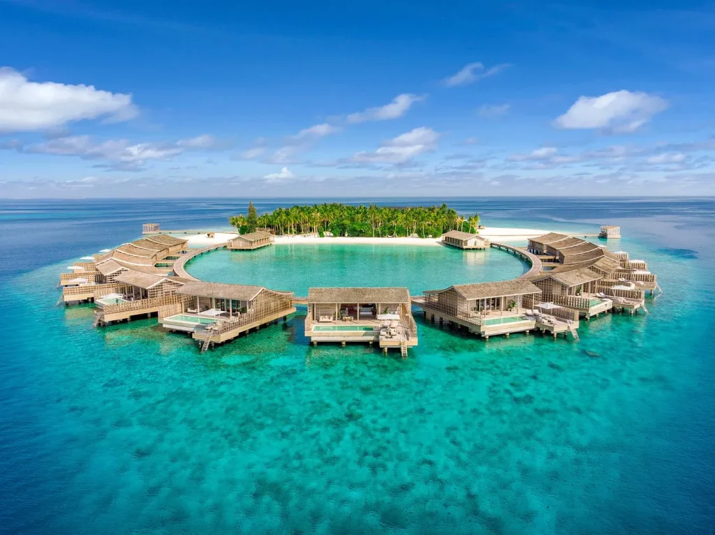 Aerial view of a circular arrangement of luxury overwater villas surrounded by turquoise ocean water and a tropical island in the Maldives.