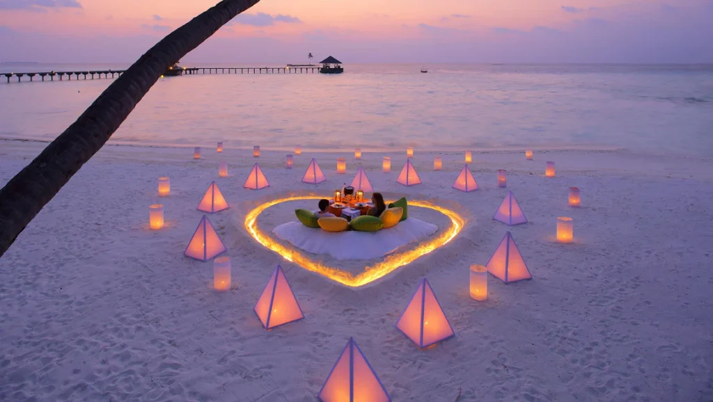 A romantic candlelit dinner for a couple inside a heart-shaped sand arrangement on a luxury Maldives beach at sunset.
