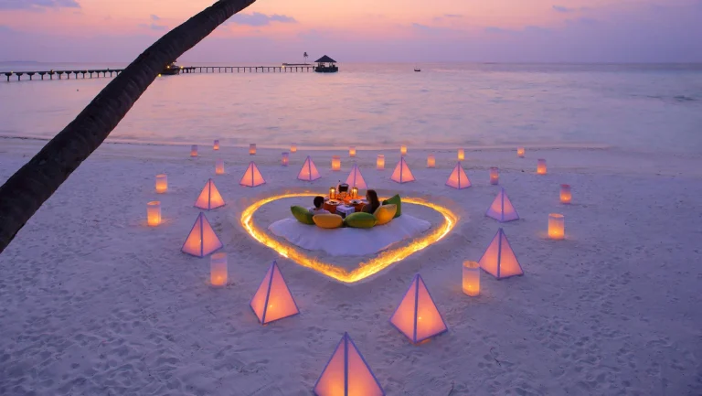 A romantic candlelit dinner for a couple inside a heart-shaped sand arrangement on a luxury Maldives beach at sunset.
