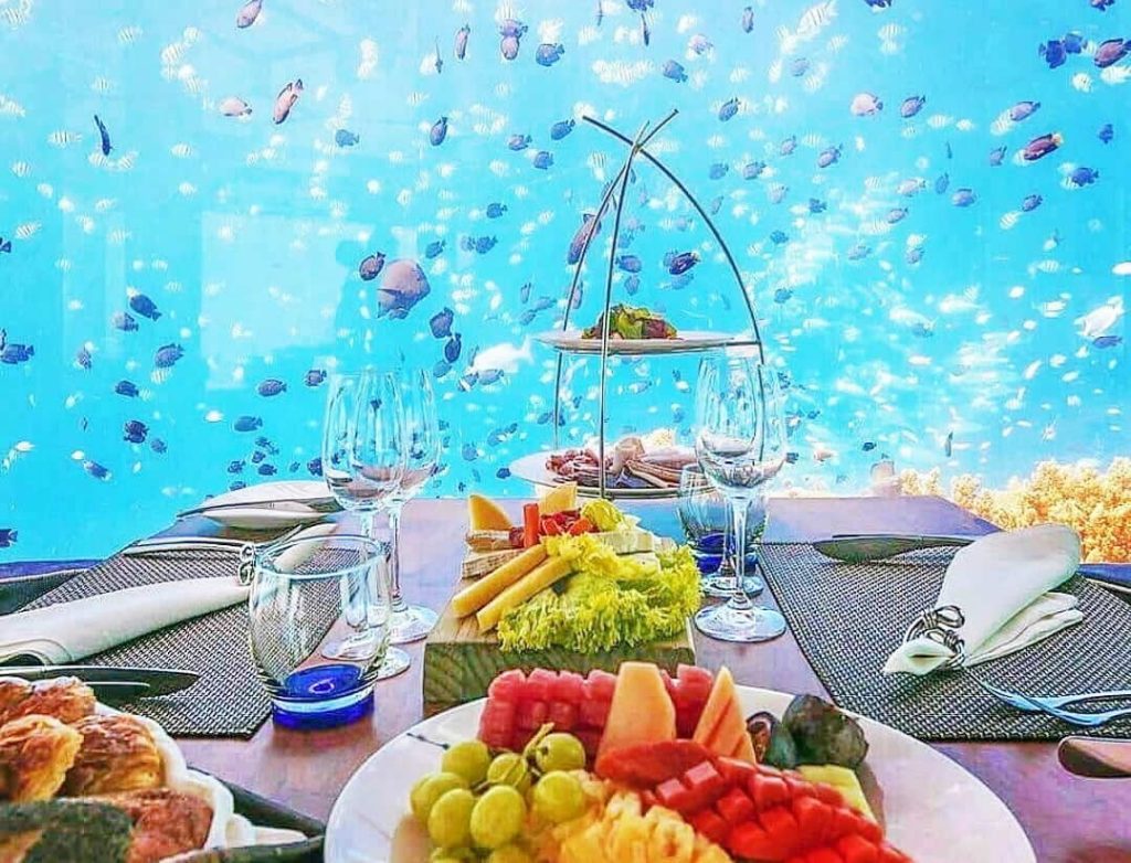 A luxury breakfast spread with fresh fruits and pastries on a wooden table inside an underwater restaurant in Lhaviyani Atoll, Maldives, with blue ocean water and tropical fish in the background.