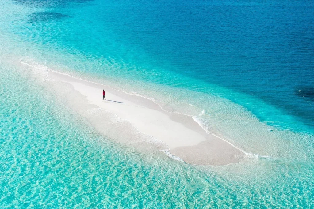 Aerial view of a lone traveler walking on a pristine white sandbank surrounded by turquoise ocean water in the Maldives.