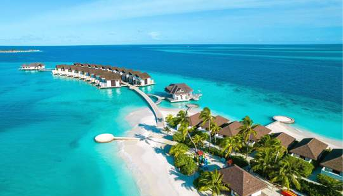 Panoramic aerial view of a lush tropical island in Vaavu Atoll, Maldives, surrounded by vibrant turquoise lagoons and deep blue ocean water under a bright sky.
