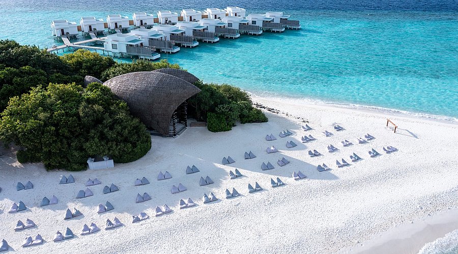 Aerial view of Dhigali Maldives luxury resort in Raa Atoll featuring overwater villas, a white sandy beach with bean bags, and a unique shell-shaped bar architecture.