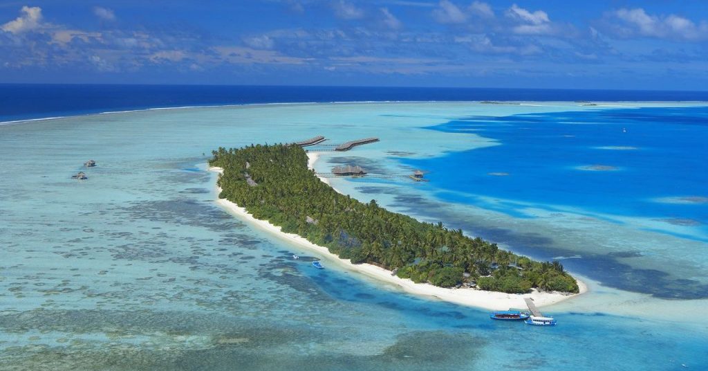 Panoramic aerial view of a lush green tropical island in Meemu Atoll, Maldives, surrounded by turquoise coral lagoons and clear blue water.