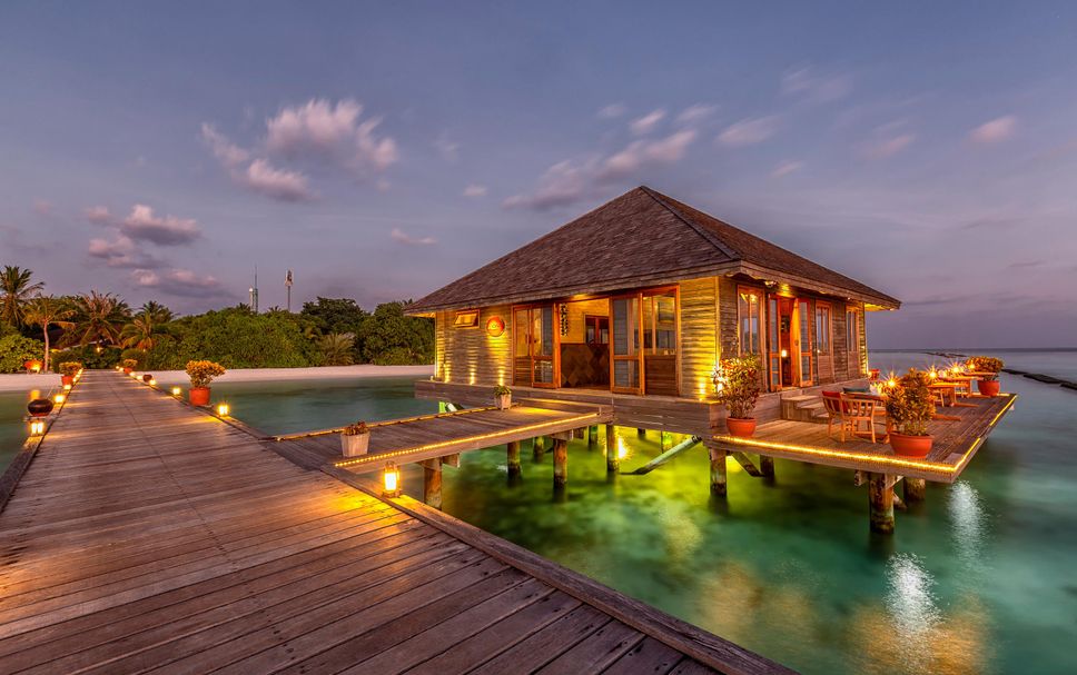 A luxury wooden overwater villa at Komandoo Island Resort in Lhaviyani Atoll, Maldives, illuminated at sunset with a wooden walkway.