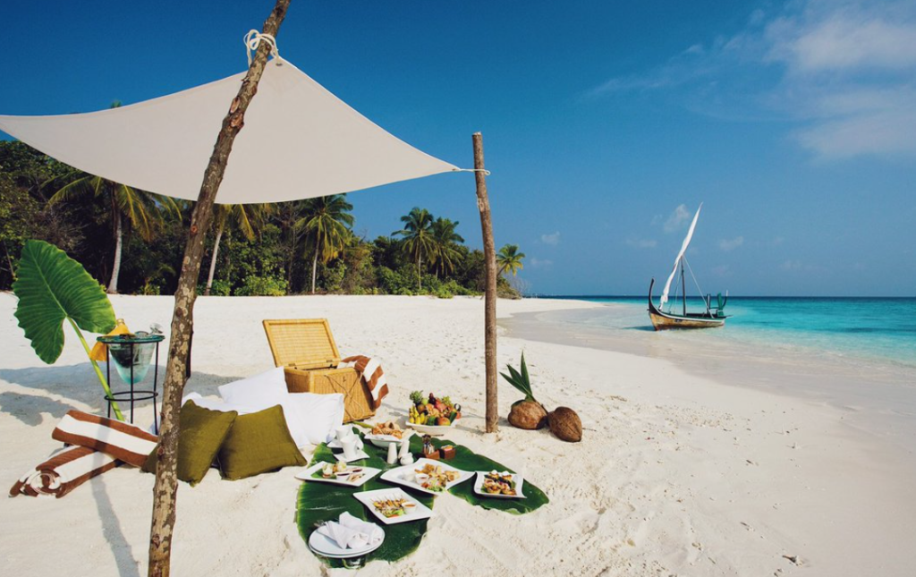 A luxury sandbank picnic setup on a white sand beach in Dhaalu Atoll, Maldives, featuring a private lunch on banana leaves, cushions, and a traditional dhoni boat in the turquoise water.