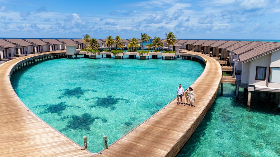 Aerial view of luxury overwater villas at Atmosphere Kanifushi resort in Lhaviyani Atoll, Maldives, featuring a family walking on a wooden boardwalk over turquoise water.