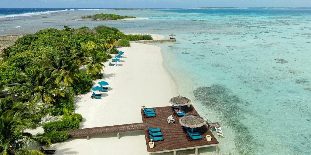 Aerial view of a luxury resort beach in Addu Atoll, Maldives, featuring a white sand shoreline, a private wooden sun deck with blue lounge chairs over turquoise lagoon water, and lush palm trees.