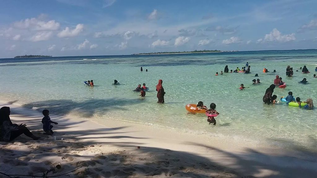 Crowded white sand beach with turquoise lagoon and families swimming, Addu Atoll, Maldives. Local island life.