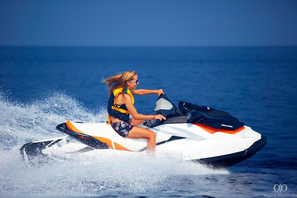 A woman riding a white and orange Sea-Doo jet ski across the deep blue waters of Gaafu Dhaalu Atoll in the Maldives, creating a large water splash.