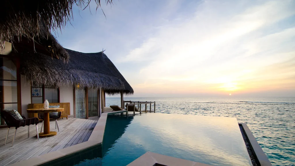 A luxury overwater villa with a private infinity pool and thatched roof at OZEN Reserve Bolifushi Maldives, showcasing a beautiful sunset over the Indian Ocean.