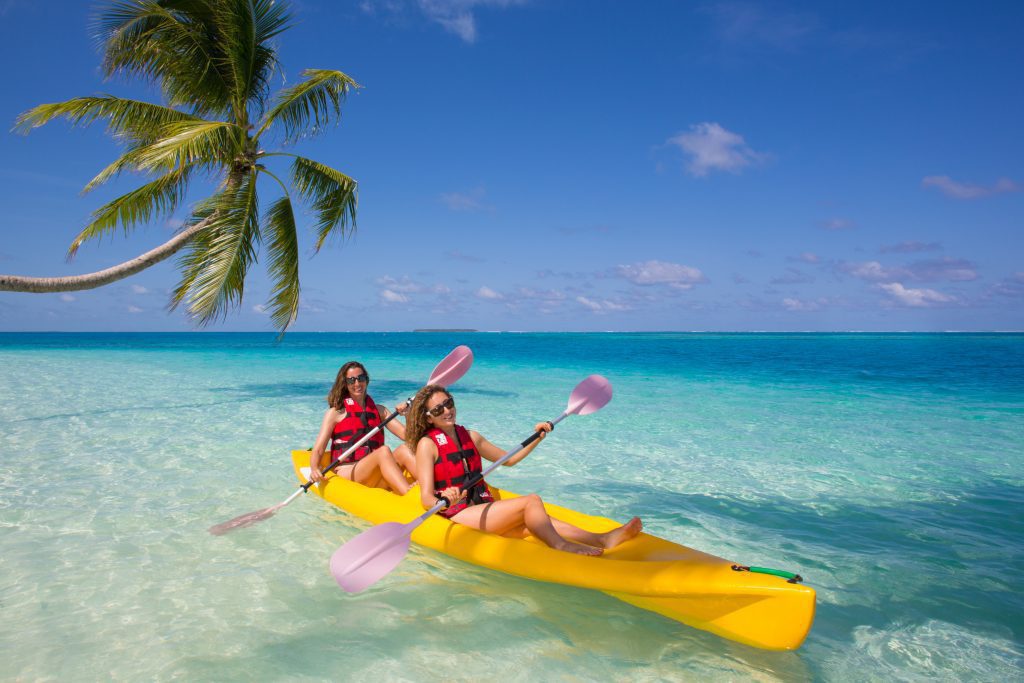 Two women in life jackets paddling a yellow tandem kayak in the turquoise shallow waters of a tropical beach in Gaafu Dhaalu Atoll, Maldives, under a palm tree.