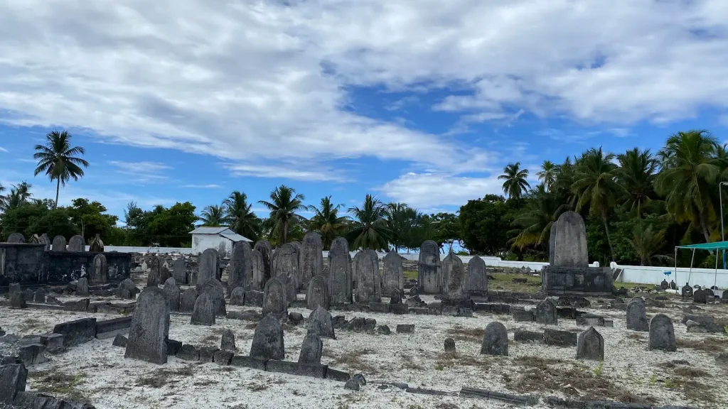A historical cemetery with numerous upright dark grey coral stone grave markers on sandy ground in Hithadhoo island, Addu Atoll, Maldives. Behind the graveyard are palm trees and dense greenery under a partly cloudy sky.