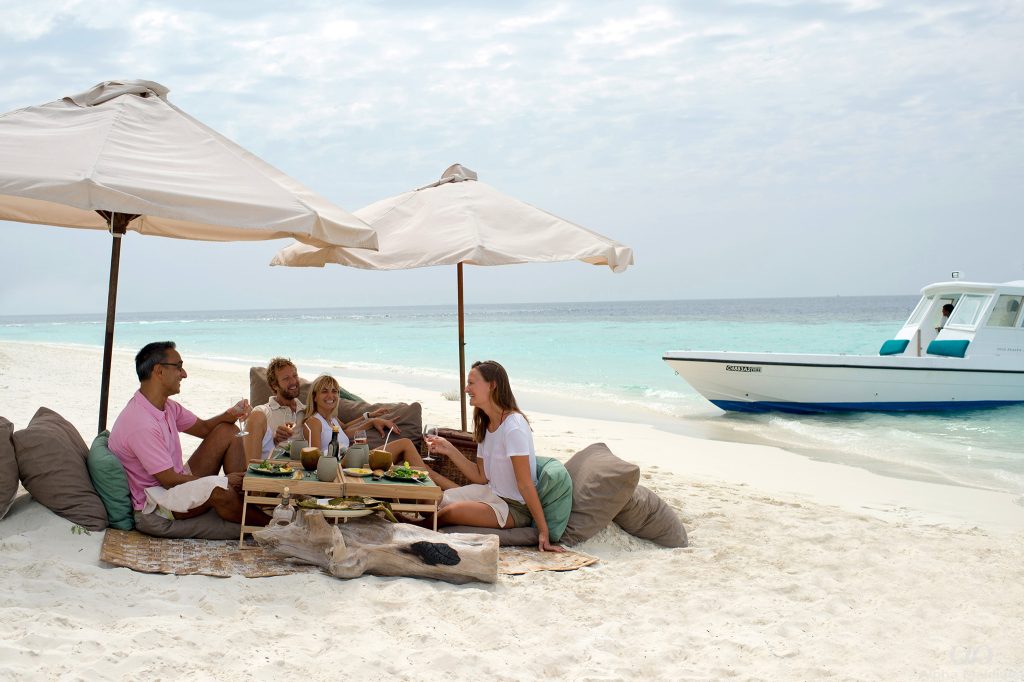 A group of travelers enjoying a private luxury sandbank picnic with gourmet food and wine on a white sand beach in Gaafu Dhaalu Atoll, Maldives.
