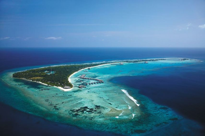 An aerial, bird's-eye photograph of an exclusive luxury island resort in Addu Atoll, Maldives. The image shows the main lush green island, its surrounding vibrant turquoise coral lagoons, multiple strings of overwater bungalows extending into the water, and the deep blue Indian Ocean under a clear sky.