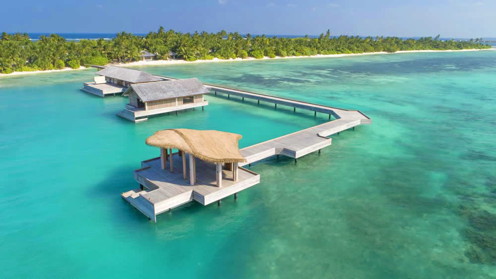 An aerial view of a luxury Maldivian resort overwater villa and boardwalk structure in the crystal clear turquoise waters of the Gaafu Dhaalu Atoll, featuring traditional thatched-roof architecture and views of a lush tropical island under a clear sky.