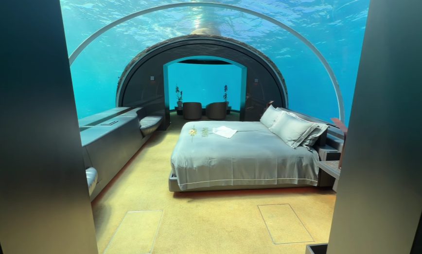Down into the underwater room goes via something labeled an escape hatch - just a straight drop, sixteen feet below sea level in the Indian Ocean. One step takes you out of polished floors and soft lighting, then suddenly walls vanish behind sweeping curves of clear plastic. Outside each curve, coral stretches wide, alive, moving slow. Space feels tight yet open at once, like stepping under water without getting wet.
Down below, things glide past like inside one of those underwater tunnels at an aquarium. Only difference being, no plastic barriers get in the way. Instead, just clear glass holds back nothing - only real sea beyond. Creatures drift over head exactly as they would if you were submerged. Light dances through surface ripples without filters or edits. What you see happens right then, shaped by tides, not timers.
Out here, where everything feels scattered, a bed sits quiet. It just appears, unplanned, between pieces of chaos. Not much explains it, yet it stays - still, low, waiting.
The Bedroom
Down below, the sleeping area stretches across the entire width of the space. A bed covered in deep ocean-colored fabric sits centered, its shade matching the watery world outside like it was meant to be there. Nearby, everyday items - a phone, places to plug in devices, electrical sockets - nestle quietly beside the mattress, easy to reach but not obvious.
Wrapped around every edge of the room, curtains can block out all light when needed. Yet those who travel here rarely pull them shut. Night reveals what lies outside - soft glows from sea creatures drifting past the windows - and few choose to miss that view. The dark water pulses gently, alive behind transparent walls, a sight most leave uncovered.
Around seven hundred square feet make up the space below the waterline. This entire area forms one connected layout beneath the surface.
Flaws exist here, yet honesty means noting each one. Stains mark the carpet, something guests likely wouldn’t anticipate at eighteen thousand dollars per night - a fix that probably belonged to pre-arrival checks. Upstairs includes extras, so finding the minibar totally bare seems odd instead. Just an espresso machine sits there, along with two bottled waters, nothing more. Not enough to walk away, though hard to ignore too.
The Sounds of Sleeping Underwater
What no image captures is the noise inside the underwater room. A steady pop echoes, like cereal soaking in a bowl - only louder, coming from everywhere at once. Most probably, the plastic walls react to tiny shifts in water push around them. Once recognized, it does not scare, yet never stops. Deep down, without thinking, you feel it: lying under tons of sea.
Rest arrives, though barely. That does not mean it fails. Just how things unfold here.
Safety and the Escape Hatch
Should anyone start thinking about emergencies, here's what matters: a bent rod sits close to the coffee maker. To open the exit overhead, grab that pole - it unlatches the way out. Nothing fancy powers it; just muscle and motion get you up. From within the room, that door is the sole sign of life beyond underwater walls. Spotting it once makes later glances easier, though most will wish they could forget its purpose entirely.
The Underwater Bathroom
Down below, the bathroom earns special mention - mainly due to housing something best called the planet’s lone loo beneath the sea. Two sinks face straight toward coral formations just outside. Behind clear panels, the shower lets you stand under clean water while saltwater stretches in every direction, full of swimming shapes. Few guest moments feel quite so dreamlike, tucked inside a home yet staring into open ocean life.
Down below, the underwater floor wraps up with a spacious closet for clothes, featuring a robe cut like a kimono, a secure box for valuables, along with a high-end hair dryer made by Dyson. On the flip side, what you get to wear on your feet stays surprisingly basic - easily the weakest part of an otherwise solid setup.